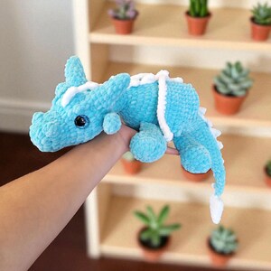 K&ouml;nnte beinhalten: Handgefertigtes, hellblaues H&auml;keldrachen-Pl&uuml;schtier mit wei&szlig;en Akzenten. Der Drache hat gro&szlig;e, glitzernde schwarze Augen und einen langen Schwanz. Das Pl&uuml;schtier ist weich und kuschelig, perfekt f&uuml;r ein Kinderzimmer oder als einzigartiges Geschenk.