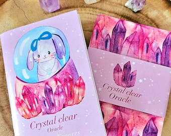 May include: A purple and pink crystal clear oracle deck with a white bunny in a snow globe on the front cover. The deck is titled "Crystal Clear Oracle" by Susana Tavares.