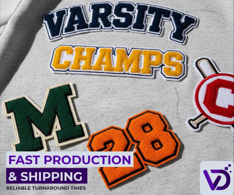 以下が含まれることがあります： 紺色と金色で「VARSITY」と「CHAMPS」の文字、緑色の「M」、オレンジ色の「28」など、さまざまな刺繍パッチのクローズアップ。野球をテーマにしたパッチも表示されています。パッチはグレーの生地にあります。