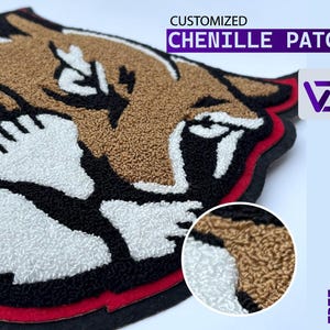 May include: A chenille patch in the shape of a roaring cougar head. The patch features brown, black, white, and red colors. The text "CUSTOMIZED CHENILLE PATCH" is displayed above the patch. The patch has a textured, raised design.