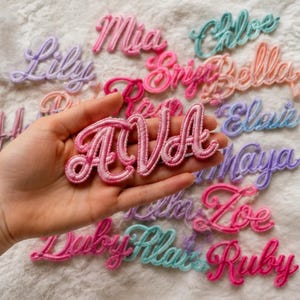 Pode incluir: Uma coleção de emblemas bordados com nomes em várias cores pastel e vivas, incluindo rosa, roxo, azul e pêssego. Cada emblema apresenta uma fonte cursiva. Um emblema rosa com o nome AVA é segurado em primeiro plano.