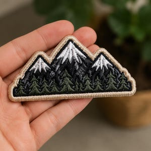 May include: An embroidered patch featuring a mountain range with white snow-capped peaks, dark gray rocky slopes, and a dense forest of green pine trees at the base. The entire design is outlined with a thick, light beige stitched border.