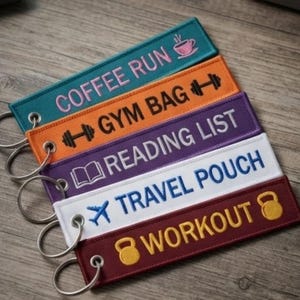 Könnte beinhalten: Fünf rechteckige Stoffschlüsselanhänger mit Metallringen. Das türkise Etikett zeigt COFFEE RUN mit einem Tassensymbol. Das orangefarbene zeigt GYM BAG mit einer Hantel. Das lila Etikett zeigt READING LIST mit einem Buch. Das weiße zeigt TRAVEL POUCH mit einem Flugzeug. Das kastanienbraune zeigt WORKOUT mit einer Kettlebell.