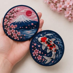 May include: Two circular embroidered patches featuring detailed thread work. One patch depicts a snow-capped mountain with a red sun and pink cherry blossoms. The second patch shows a white, red, and black koi fish swimming in blue water.