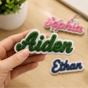 May include: A custom embroidered name patch featuring the name Aiden in dark green cursive thread on a white felt background. Additional patches with the names Sophia in pink and Ethan in navy blue are visible in the background.