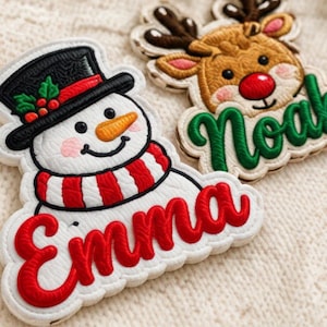 May include: Two embroidered patches featuring holiday characters. One patch shows a snowman wearing a black top hat and red-striped scarf with the name Emma in red. The other patch shows a reindeer with a red nose and the name Noah in green.