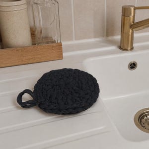 May include: A black, round, crocheted dish scrubby with a loop handle. The scrubby is sitting on the edge of a white sink. A gold faucet is visible in the background.
