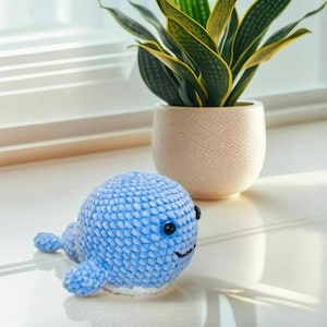 Peut inclure: Un jouet baleine crocheté bleu clair avec des yeux noirs et une bouche souriante. La baleine en peluche a de petites nageoires et un ventre blanc. Une plante en pot avec des feuilles vertes et jaunes est en arrière-plan.