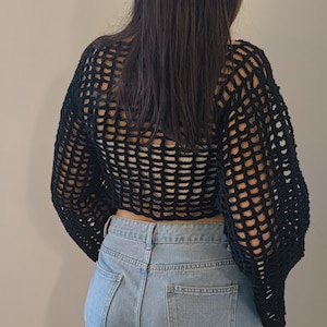 Peut inclure: Un haut court noir en maille ajourée à manches longues. Le haut est porté avec un jean en denim bleu clair. L'image est prise de dos, montrant la texture du haut et la coupe du jean.