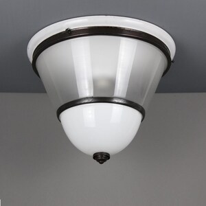 Op de afbeelding: Een witte en matglazen plafondlamp met een donkere metalen rand. De lamp heeft een afgeronde bodem en een conische, matglazen kap. De armatuur is op een grijs plafond gemonteerd.