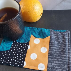 Könnte beinhalten: Ein Patchwork-Untersetzer mit Stoff in Türkis, schwarzen Punkten, orangefarbenen Punkten und grauen Chevron-Muster. Eine Tasse Tee mit Zimtstange und eine ganze Orange liegen neben dem Untersetzer.