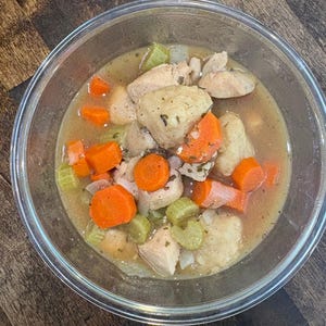 Puede incluir: Un cuenco de vidrio transparente lleno de estofado de pollo. El estofado contiene trozos de pollo cocido, rodajas de zanahoria naranja, trozos de apio verde y un caldo marrón claro. El cuenco está sobre una superficie de madera.