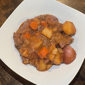 以下が含まれることがあります： 白い正方形の皿に盛られたビーフシチュー。シチューには、肉、ジャガイモ、ニンジンが濃厚な茶色のグレービーソースで入っています。ボリュームたっぷりの食事です。