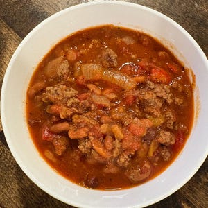 Puede incluir: Un cuenco blanco lleno de chili abundante. El chili es de color rojo intenso y contiene carne picada, frijoles, tomates, cebollas y pimientos. El cuenco está sobre una superficie de madera oscura.