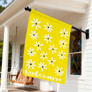 Könnte beinhalten: Eine leuchtend gelbe, rechteckige Gartenflagge mit einem wiederkehrenden Muster aus weißen Gänseblümchen mit schwarzen Zentren. Das Wort "welcome" ist in weißer Schreibschrift am unteren Rand der Flagge aufgedruckt, die an einer schwarzen Metallstange hängt.