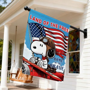 May include: A decorative garden flag featuring a cartoon dog wearing aviator goggles and a scarf, piloting a red biplane. The background displays a waving American flag with the text LAND OF THE FREE and HOME OF THE BRAVE in bold lettering.