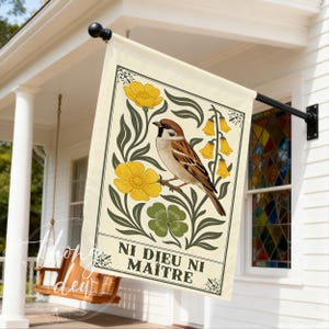 Könnte beinhalten: Eine cremefarbene Gartenflagge mit einer detaillierten Illustration eines Spatzen, der auf einem Ast sitzt, umgeben von gelben Blumen, grünen Blättern und einem vierblättrigen Kleeblatt. Die Flagge trägt den Text "NI DIEU NI MAÎTRE" in Schwarz.