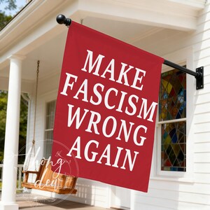 Könnte beinhalten: Eine rechteckige rote Flagge, die an einem schwarzen Metallmast hängt. Die Flagge zeigt den weißen Text "MAKE FASCISM WRONG AGAIN" in einer fetten serifenlosen Schrift. Die Flagge ist vor der Außenseite eines weißen Hauses angebracht.