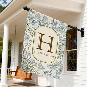 May include: A rectangular garden flag featuring a cream background with a blue toile-style pattern of a bridge, house, flowers, and ducks. A central oval frame contains a large brown letter H above the text THE HARRISONS in brown lettering.