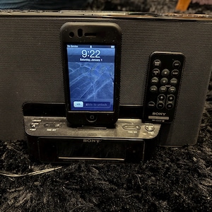 May include: Black Sony speaker system with a docked iPhone displaying the time 9:22 on January 1st. A black remote control is to the right. The speaker system sits on a black, textured surface.