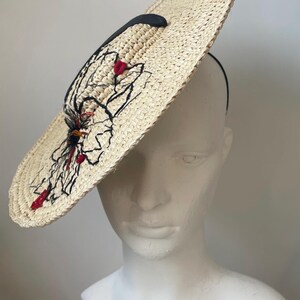 May include: A wide-brimmed straw hat with a black and red embroidered design. The hat has a black band and is displayed on a white mannequin head. The hat's design includes abstract shapes and floral elements.