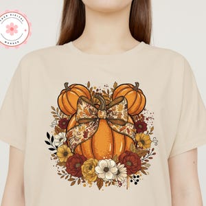 May include: Beige t-shirt with a fall-themed graphic. The design features an orange pumpkin with a decorative bow, surrounded by pumpkins, flowers, and foliage in orange, yellow, and burgundy. The shirt has a round neckline.