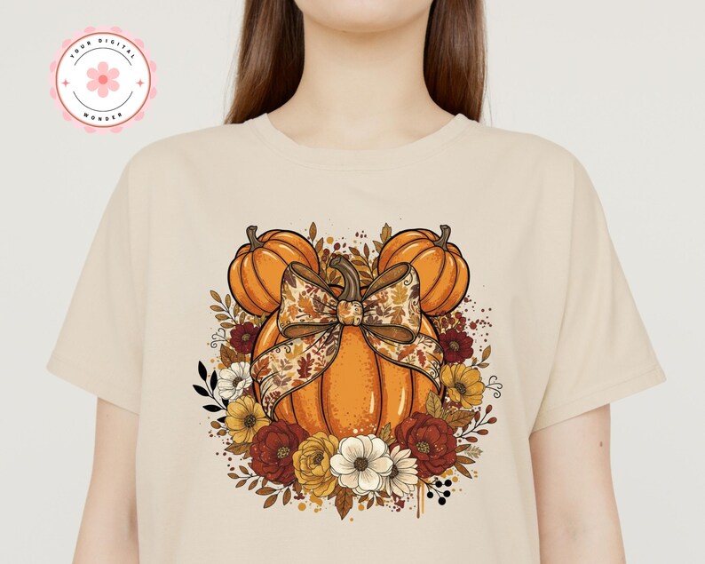 May include: Beige t-shirt with a fall-themed graphic. The design features an orange pumpkin with a decorative bow, surrounded by pumpkins, flowers, and foliage in orange, yellow, and burgundy. The shirt has a round neckline.