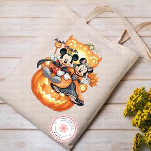 K&ouml;nnte beinhalten: Beige Tragetasche mit einem Halloween-Design mit Mickey und Minnie Mouse in Kost&uuml;men, die auf einem orangefarbenen K&uuml;rbis sitzen. Die Tasche hat lange Henkel und ein kleines, rundes Logo unten.