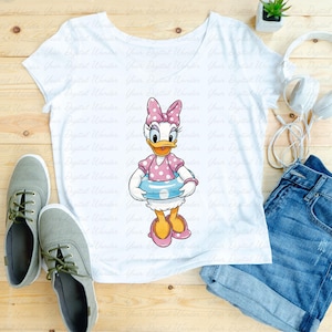 May include: White t-shirt featuring a cartoon image of Daisy Duck wearing a pink polka dot bow, dress, and shoes, and holding a blue and white inner tube. The shirt is paired with denim shorts and gray sneakers.