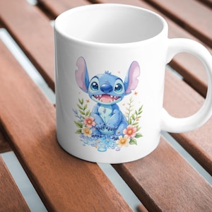 May include: White ceramic mug featuring a blue cartoon character with large ears and a wide smile, surrounded by colorful flowers and foliage. The character is sitting on a bed of blue water droplets. The mug has a curved handle.