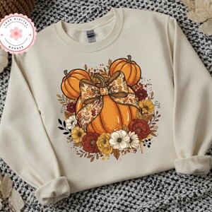 May include: A cream-coloured sweatshirt with a large orange pumpkin design, a decorative bow, and autumn flowers. The design includes smaller pumpkins and a floral arrangement in orange, red, and yellow. The sweatshirt has long sleeves and a crew neck.