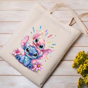 May include: Beige canvas tote bag featuring a colorful illustration of two cartoon aliens, one blue and one pink, surrounded by flowers and colorful paint splatters. The bag has two beige handles.
