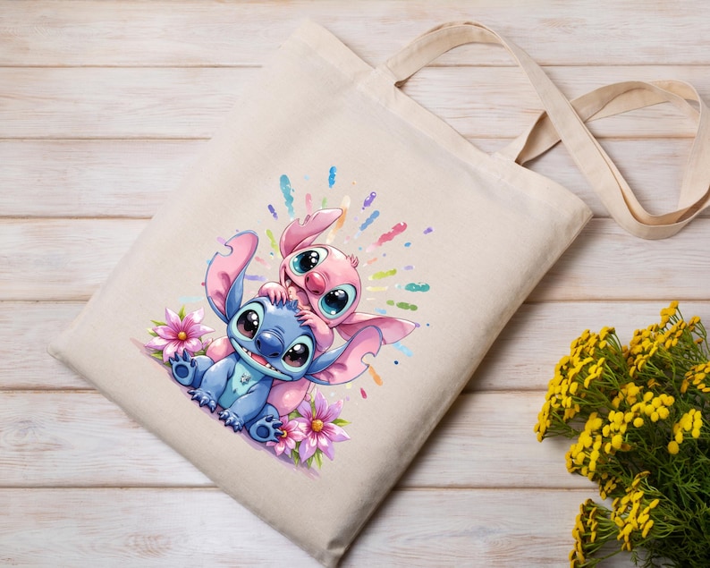 May include: Beige canvas tote bag featuring a colorful illustration of two cartoon aliens, one blue and one pink, surrounded by flowers and colorful paint splatters. The bag has two beige handles.