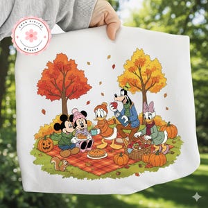 May include: White fabric featuring a fall picnic scene with Mickey, Minnie, Donald, Goofy, and Daisy. The characters are gathered around a picnic blanket with pumpkins, pie, and a basket of food. Autumn trees and falling leaves complete the scene.