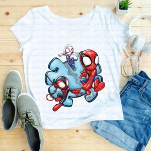 May include: White t-shirt with a vibrant graphic of Spider-Man and two other Spider-Verse characters. The shirt is styled with denim shorts, grey trainers, and white headphones on a wooden surface.