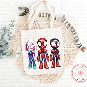 May include: A cream canvas tote bag displaying three cartoon superheroes: Spider-Gwen, Spider-Man, and Miles Morales. The superheroes are in their iconic costumes, with bright colours and strong outlines. The bag rests on a neutral-toned woven surface.