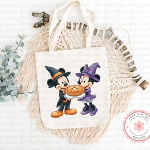 K&ouml;nnte beinhalten: Eine wei&szlig;e Canvas-Tragetasche mit einem Halloween-Motiv von Mickey und Minnie Mouse. Mickey tr&auml;gt einen schwarzen Hexenhut und h&auml;lt einen Kuchen. Minnie tr&auml;gt einen lila Hexenhut und ein Kleid. Die Tragetasche hat cremefarbene Griffe.
