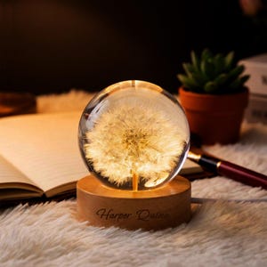 May include: A clear glass sphere with a dandelion design, resting on a wooden base. The base is engraved with the text "Harper Quinn". The sphere is illuminated, creating a warm glow. A pen, a book, and a small potted plant are in the background.