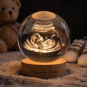 Op de afbeelding: Een heldere glazen bol met een verlichte echografie van een baby. De bol staat op een houten voet met de naam "Finnian Cole" gegraveerd. Een teddybeer en babyschoentjes zijn op de achtergrond te zien.