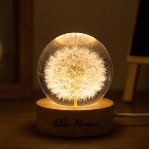 May include: A glowing glass sphere with a dandelion design, resting on a wooden base. The base is engraved with the name "Ella Vance". The sphere emits a warm, inviting light, creating a decorative and unique lighting piece.