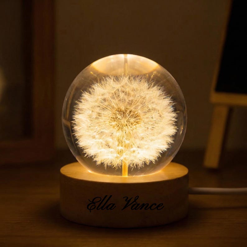 May include: A glowing glass sphere with a dandelion design, resting on a wooden base. The base is engraved with the name "Ella Vance". The sphere emits a warm, inviting light, creating a decorative and unique lighting piece.