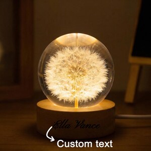 May include: A glowing glass sphere with a dandelion design, resting on a wooden base. The base has the text "Ella Vance" and "Custom text" engraved. The sphere emits a warm, inviting light, perfect for a nightstand or desk.