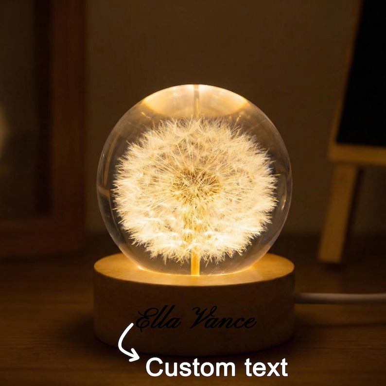 May include: A glowing glass sphere with a dandelion design, resting on a wooden base. The base has the text "Ella Vance" and "Custom text" engraved. The sphere emits a warm, inviting light, perfect for a nightstand or desk.