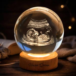 Puede incluir: Una bola de cristal transparente con una imagen de ultrasonido de un bebé en su interior, colocada sobre una base de madera iluminada desde dentro. La base tiene grabado el nombre "Harper Quinn". El fondo es oscuro con iluminación suave.