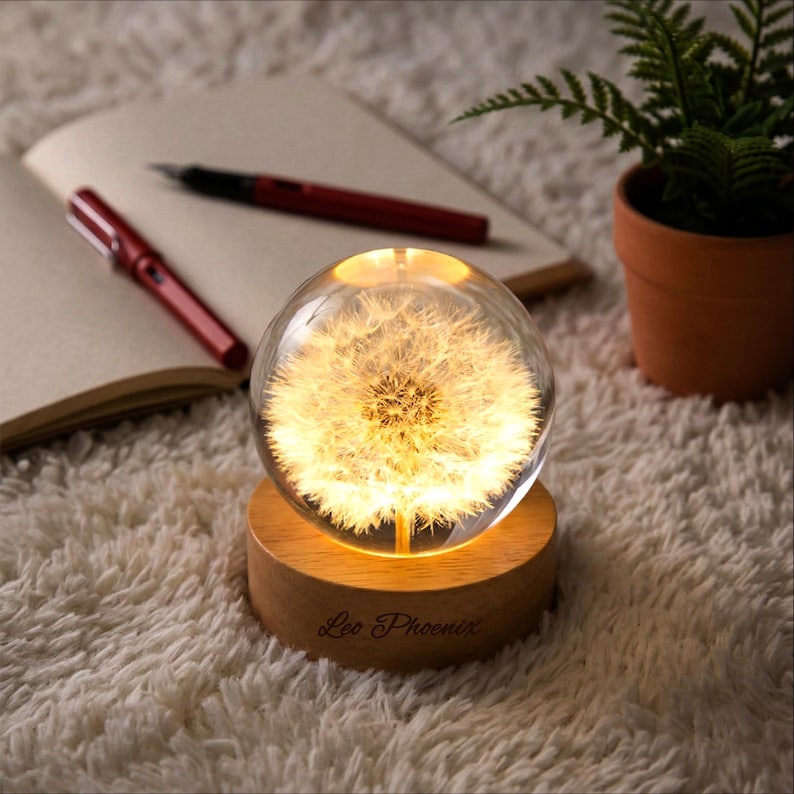 May include: A clear glass sphere with a dandelion design, illuminated from within, sits on a wooden base. The base has the text "Leo Phoenix" engraved on it. The sphere is on a white, fluffy surface, with a notebook and pens in the background.