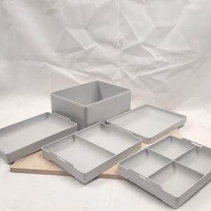 May include: A set of light gray storage containers, including a large square box and several trays of varying sizes. Some trays are divided into compartments. The containers are arranged on a light-colored wooden surface.