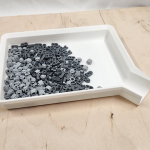 May include: A white plastic tray filled with a collection of small, gray and black building block pieces. The tray has a sloped side for easy access. The blocks are of various shapes and sizes, suitable for construction projects.