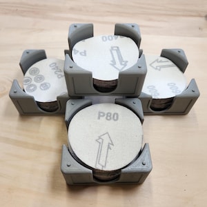 以下が含まれることがあります： 研磨ディスク用の灰色のプラスチック製収納容器が4つあります。各容器には、P80、P120、P240、P400という異なる粒度の研磨ディスクが積み重ねられています。研磨ディスクは白地に黒文字で、上向き矢印が描かれています。