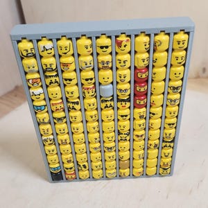 May include: A gray wooden display case filled with yellow Lego heads. The heads have various facial expressions, including sunglasses, smiles, and frowns. Some heads are red or blue. The case is rectangular and has multiple compartments.