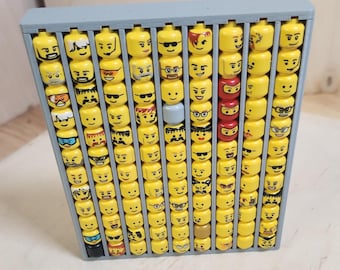 May include: A grey wooden display case filled with yellow Lego heads. The heads have various facial expressions, including sunglasses, smiles, and frowns. Some heads are red or blue. The case is rectangular and has multiple compartments.